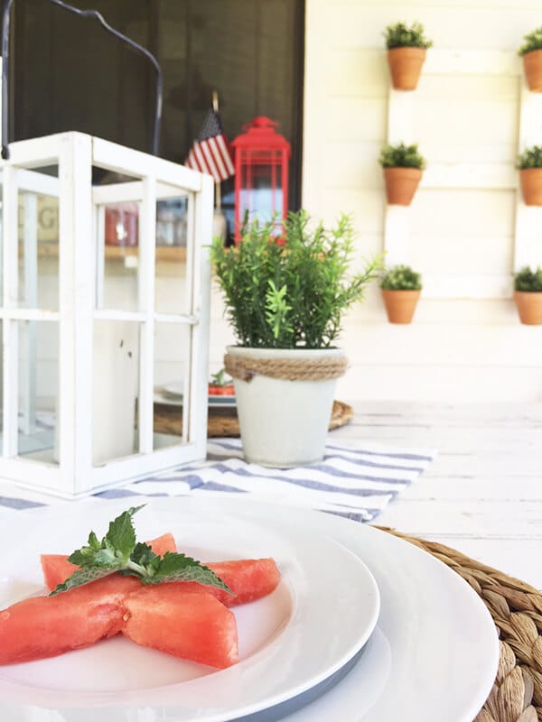 Farmhouse style patriotic outdoor tablescape for the 4th of July. Striped fabric, watermelon, seagrass placemats, and red, white and blue, all on a chippy farmhouse table.