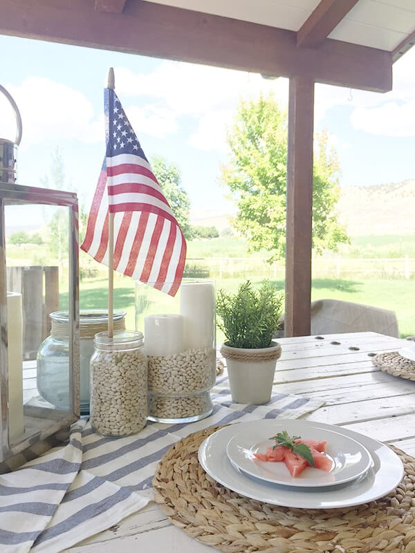Farmhouse style patriotic outdoor tablescape for the 4th of July. Striped fabric, seagrass placemats, and red, white and blue, all on a chippy farmhouse table.