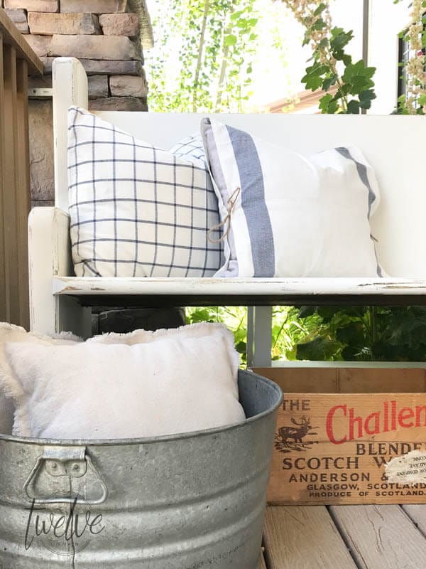 This incredible farmhouse style church pew is the perfect addition to this farmhouse porch! You have to see what it looked like before. With the addition of farmhouse pillows, crates, baskets, and galvanized tubs, this space is perfection.