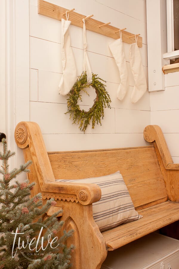 Create beautiful farmhouse Christmas entry decor with a vintage church pew, simple white Christmas stockings, a boxwood wreath, grainsack pillows, and a fresh blue spruce Christmas tree.