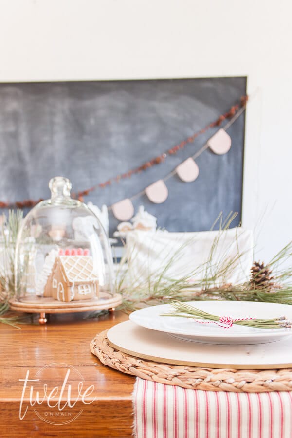 Mini gingerbread houses in glass cloches on the table, red and white Christmas garland along the chalkboard, vintage pottery, Rae Dunn Clay pottery, and pine boughs.