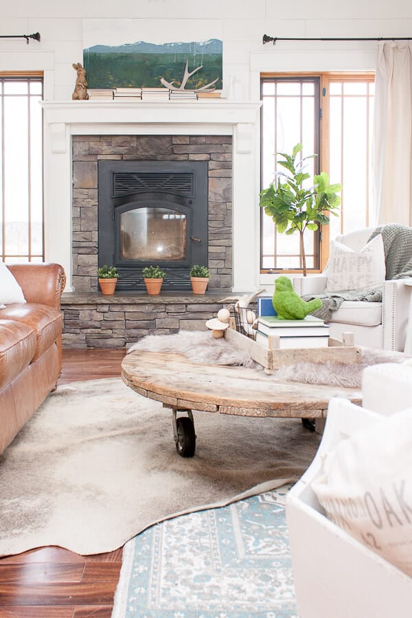 Cozy farmhouse living room with drop cloth curtains, farmhouse artwork, hardwood floors, and a stacked stone fireplace.
